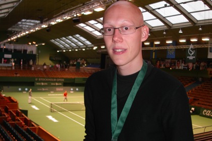 Johan Porsborn står högt upp på läktaren med tennisplan i bakgrunden. Han bär glasögon, svart tröja och är skallig.