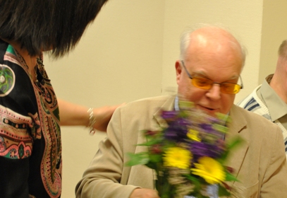 Göran Ringblom har tunt vitt hår och glasögon. Han avtackas med blommor.