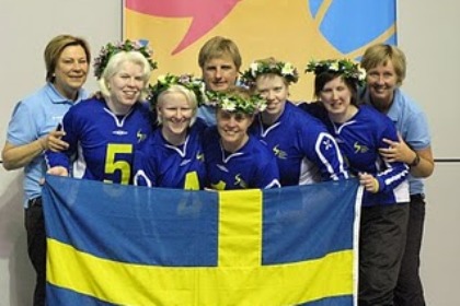 Svenska damlaget i goalball med blommar i håret och deras tränare håller upp en stor svensk flagga.