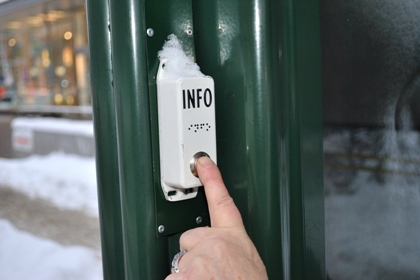 En prator, vit och stor som en liten tändsticksask, med texten info i svart och punkt och med en metallknapp. Pratorn är fäst på busskurens gröna metallram och vi ser ett finger trycka på knappen.