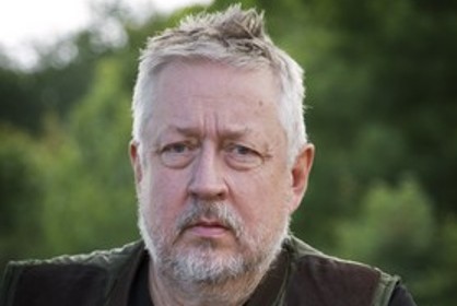 Leif GW Persson har runt ansikte, rufsigt vitt hår och skägg. Han har en dubbelhaka och stora påsar under ögonen