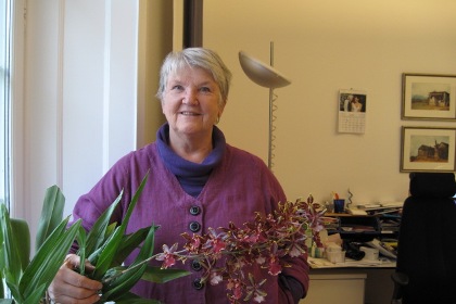 Meta Troell har grått kortklippt hår. Hon har en mörklila polo och en ljuslila linnéjacka. Framför sig har hon en blommande orkidé.