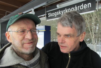 Mats Sandell har grön keps kort grått skägg och bär glasögon. Bengt Troberg bär svart jacka och har grått kortklippt hår. De står på perrongen till Skogskyrkogården