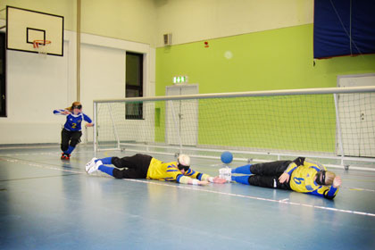 Spelare från svenska damlandslaget  i goalball tränar inför EM i Danmark. Två spelare ligger ner på marken för att blockera bollen. En tredje spelare springer fram mot planens mitt från målets ena kortsida.