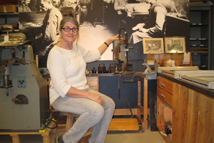 Gilda Stiby sitter på en pall vid en borstbindarbänk på Synskadades Museum. Hon har långt gråbrunt hår uppsatt i en knut och glasögon. I bakgrunden syns en stor svartvit bild från ett borstbinderi.