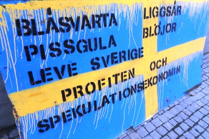 Plakat vid Occupy Stockholm-läger, föreställande svensk flagga med ett antal ord skrivna i svart text; Blåsvarta, pissgula, Liggsår, Blöjor, Leve Sverige, profiten och spekulationsekonomin.