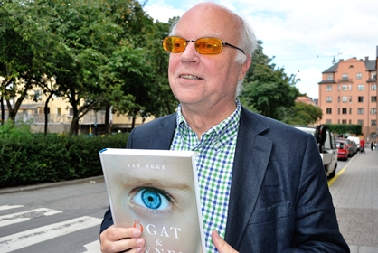 Göran Ringblom i vitt hår och orangea glasögon håller upp en bok med ett stort öga på framsidan.