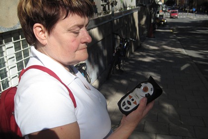 Siw Olsson står på en trottoar och håller i sin GPS . Siv har kortklippt brunt hår och en vit t-shirt. Apparaten är grå med svarta och orangea knappar.