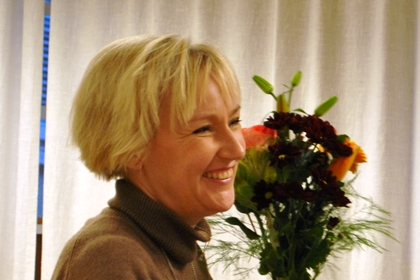 Helene Hellmark Knutsson har ganska kort blont hår och brun polotröja. Hon håller i en blomsterkvast och ler.