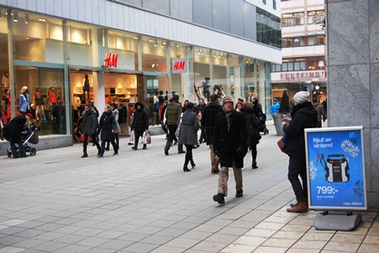 Vinterklädda stockholmare jäktar fram på sergelgatan. I Bakgrunden ses en Hennes och Mauritzbutik som skyltar med vårmodet.