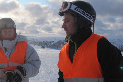 Bild på två män klädda för skidåkning, men orangea västar och fjäll i bakgrunden.