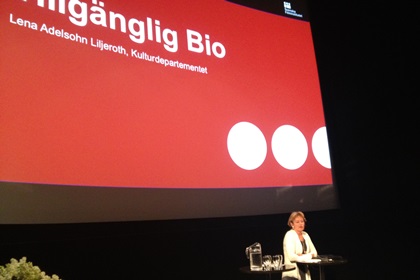 På bilden ser vi en kvinna på ett podium. I bakgrunden texten ”Tillgänglig bio” mot röd bakgrund.