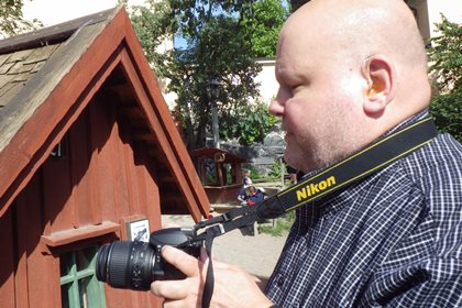 Rolf Eriksson bär en mörk smårutig skjorta och står med kamera framför ett av de små husen i lekparken. Han saknar hår på huvudet.